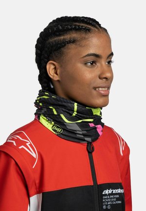 Persona con capelli intrecciati che indossa una giacca rossa e una scaldacollo nera multicolore con accenti gialli e rosa.