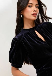 Morska modra žametna obleka z izrezom v obliki ključavne luknje, nabranimi rokavi in nabranimi detajli. Tkanina ima mehak, gladek teksturo.