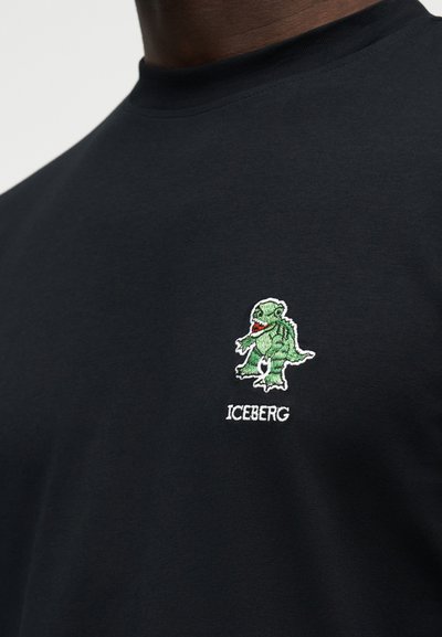 Εγγύς πλάνο μαύρου πουκάμισου με μικρή πράσινη κεντημένη δεινοσαυρο πάνω από το λευκό κείμενο «ICEBERG» στο στήθος.