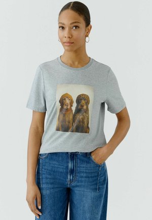 Jonge vrouw met creolen die een grijs T-shirt draagt met een print van twee honden, waarvan één met een gebreide muts, gecombineerd met een blauwe spijkerbroek.