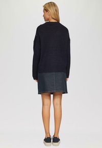 Marineblauer gestrickter Pullover mit überschnittenen Schultern, kombiniert mit einem dunklen Jeansrock. Legere Sneaker runden das Outfit ab. Weicher Stoff, lockere Passform.