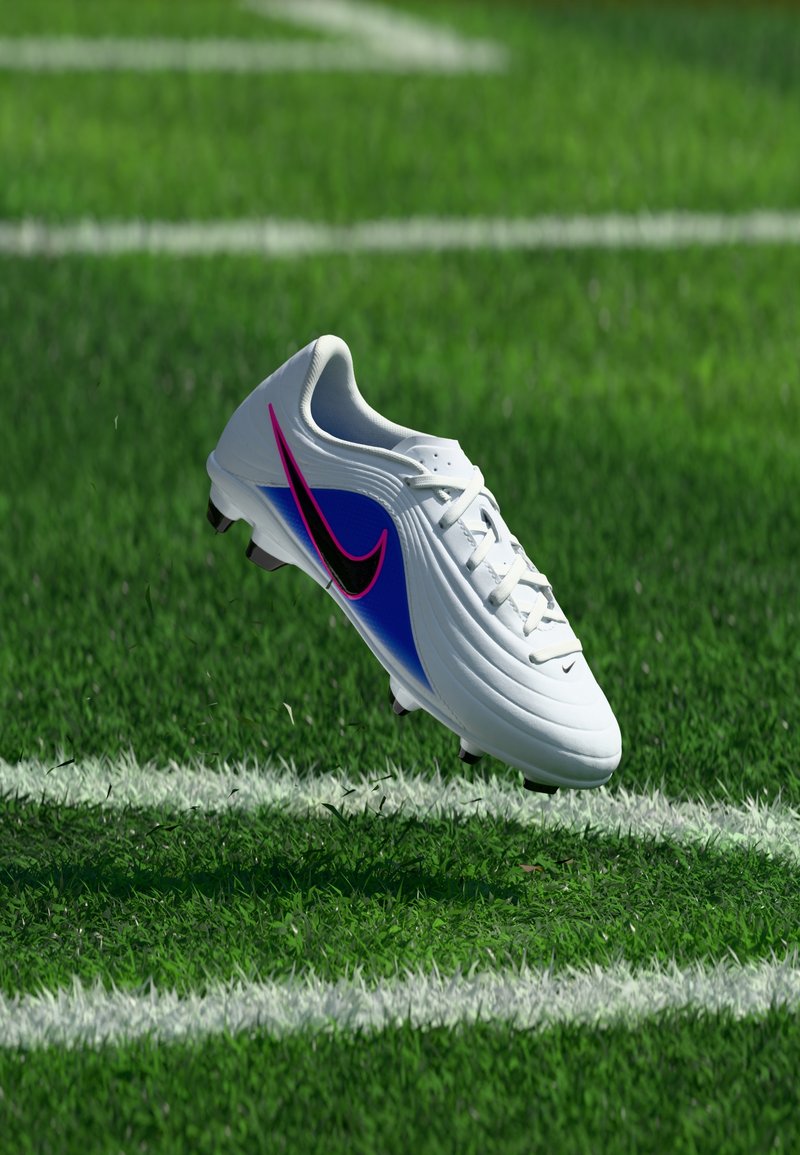 Chaussure de football blanche avec un swoosh Nike noir et rose, accent bleu, flottant au-dessus d'un terrain en gazon vert avec des lignes blanches.
