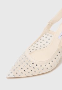 Cream-colored slingback pump with a pointed toe, embellished with silver rhinestones, and a transparent polka-dot mesh side panel.
