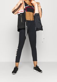 Schwarze und orange Windjacke mit gestreiften Akzenten, kombiniert mit einem schwarzen Sport-BH und figurbetonten schwarzen Jogginghosen mit einem seitlichen Muster. Schwarze Sneakers.