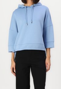 Sudadera con capucha azul de corte holgado, mangas tres cuartos, detalles de nudo en los laterales y textura suave y lisa. Combinada con pantalones negros.
