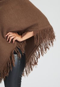 Brun vävd poncho med fransiga kanter, texturerad stickad tyg, bår över svarta byxor, som visar en lös, rektangulär form.