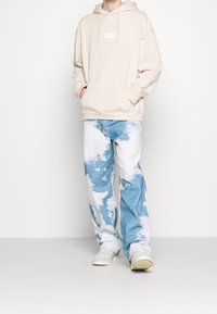 Beige-huppari, jossa etutasku ja kiristysnauhat, yhdistetty sinivalkoisiin liukuvärjättyihin farkkuihin. Malli käyttää valkoisia lenkkareita.