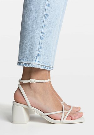 Fødder iført hvide sandaler med blokhæl og stropper, kombineret med lyseblå cropped jeans mod en ensfarvet baggrund.