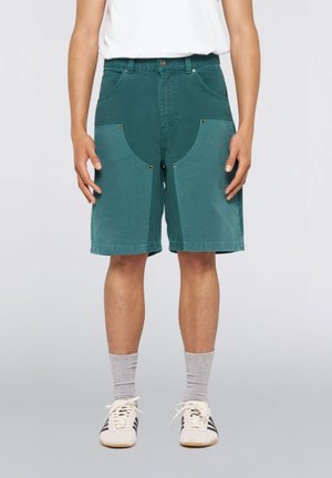 Uomo che indossa pantaloncini a due tonalità di verde fino al ginocchio con tasche anteriori, calzini grigi a coste e scarpe da ginnastica bianche con strisce nere.