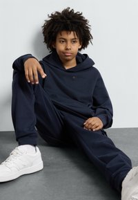 Navyblauwe hoodie en joggingset gemaakt van zachte stof, met een logo aan de voorkant. Witte sneakers met een gestructureerde zool maken de outfit compleet.