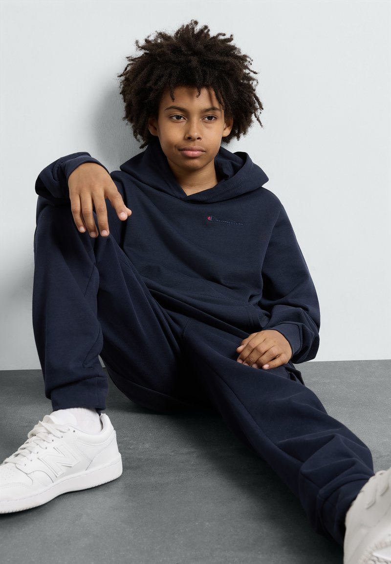 Granatowy zestaw bluzy z kapturem i spodni dresowych wykonany z miękkiego materiału, z logo z przodu. Białe sneakersy z teksturowaną podeszwą dopełniają stylizację.