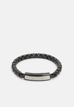 Bracciale in metallo nero con un bracciale flessibile, una placca centrale in argento con la parola "BOSS" incisa e una superficie lucidata.
