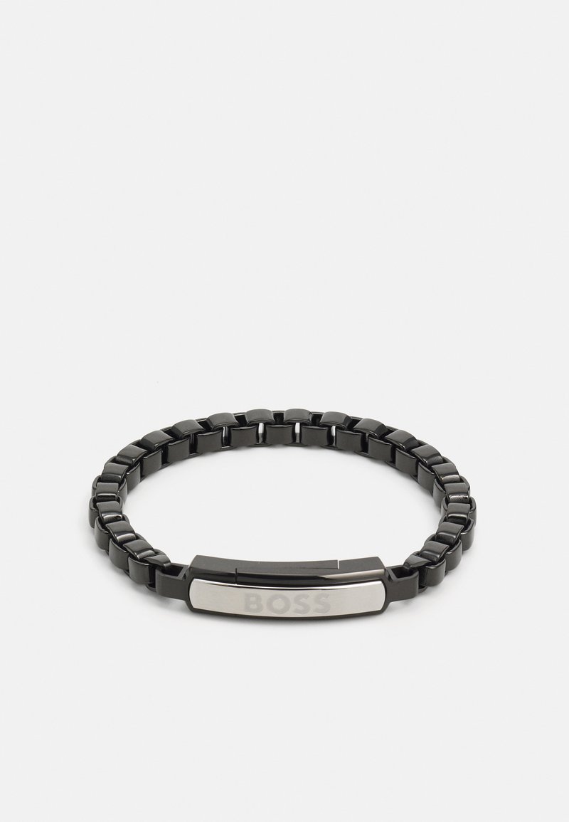 Pulsera de metal negro con una banda flexible, una placa central de plata con la palabra "BOSS" grabada y una superficie pulida.