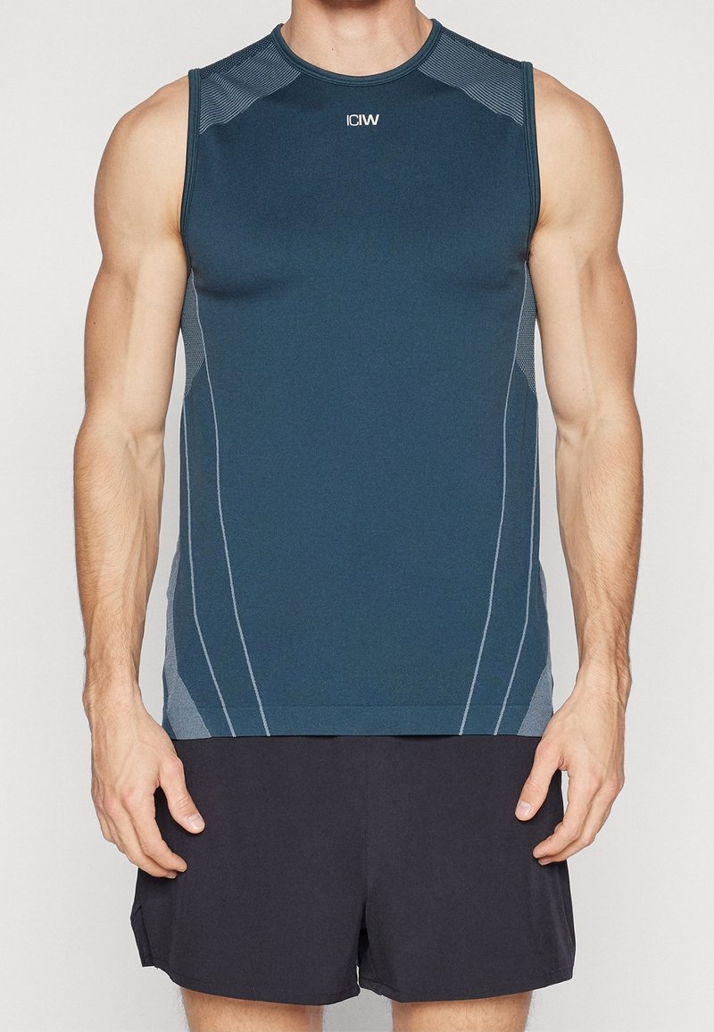 Personne musclée portant un débardeur de sport bleu avec le logo "ICW" et un short de sport noir, debout devant un fond uni.