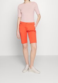 Persona con una camiseta ajustada de color rosa claro, pantalones cortos naranja brillante hasta la rodilla y zapatillas blancas, de pie en un suelo gris claro frente a una pared blanca.