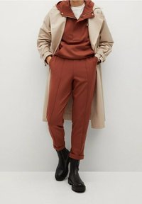 Beżowy płaszcz trencz na rdzawopomarańczowej bluzie z kapturem oraz dopasowanych, zwężanych spodniach. Czarne botki za kostkę. Gładka tkanina, minimalne zdobienia.