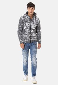 Grau bedruckter Hoodie mit Kordelzügen, der eine Mischung aus Designs zeigt. Kombiniert mit hellblauen Slim-Fit-Jeans mit Cargo-Taschen. Weiße Sneakers.