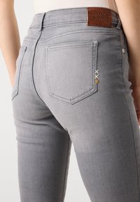 Grå denimjeans med høy midje, baklommer med trekantet form og dekorativ søm i gul og hvit. Lærmerke.