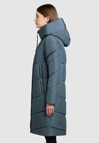 Kvinde iført en knælængde, blå polstret frakke med høj krave, hætte og lynlåslommer, der står i profil mod en ensfarvet baggrund.