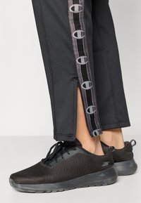 Μαύρα αθλητικά παπούτσια από πλεκτό υλικό με υφή, κλείσιμο με κορδόνια και διαφανές πάτο. Συνδυασμένα με μαύρο παντελόνι που έχει διακοσμητική ρίγα στο πλάι.