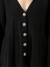 Black cardigan with a V-neck, featuring vertical pleats and silver floral buttons along the front. Textured knit fabric.