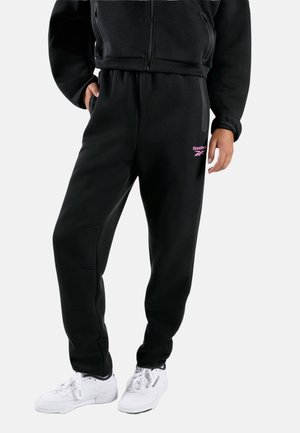 Personne portant un pantalon de survêtement noir Reebok avec un logo rose, une veste noire zippée et des baskets blanches Reebok, debout avec une main dans la poche.