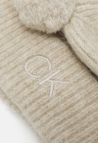 Cappello in lana lavorato a maglia beige con texture a coste, dotato di un pom-pom peloso e logo ricamato in rosa chiaro. Materiale morbido e caldo.