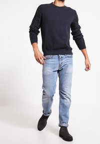 Marinblå tröja med texturerade kabelstickade ärmar, i kombination med ljus denimjeans och svarta ankelstövlar. Ren design med en avslappnad passform.