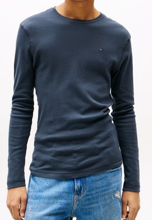 Homme portant une chemise ajustée bleu foncé à manches longues avec un petit logo sur la poitrine et un jean bleu clair usé.