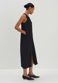 Schwarzes ärmelloses Kleid mit seitlichen Taschen, elongated Form und einem Schlitz vorne. Hergestellt aus einem glatten Stoff, kombiniert mit schwarzen spitzen Schuhen.
