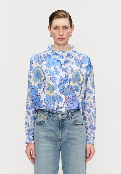 Bluse mit Blumenmuster in Blau- und Gelbtönen, mit Rüschensaumkragen und langen Ärmeln, aus leichtem Stoff gefertigt. Getragen mit hellen Jeans.
