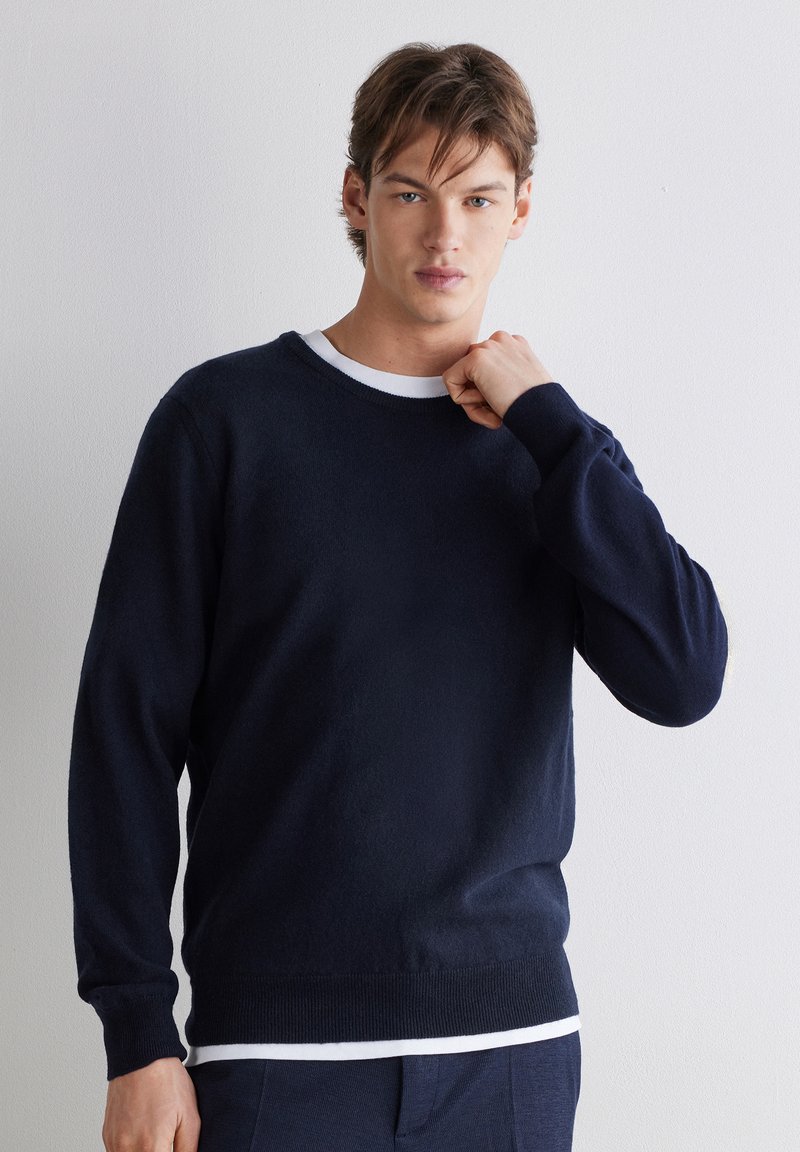 Jeune homme aux cheveux bruns courts portant un pull bleu marine foncé par-dessus une chemise blanche, debout devant un mur clair uni.