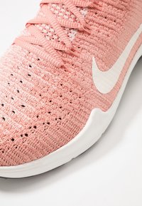 Närbild av en rosa träningsskor med en texturerad stickad överdel och vit swoosh-logotyp, som framhäver snörningarna och den ventilerande designen.