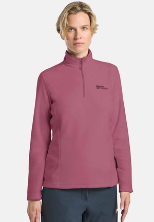 Vrouw met kort blond haar, gekleed in een mauve fleece sweatshirt met kwartrits en donkere broek, staande tegen een effen witte achtergrond.