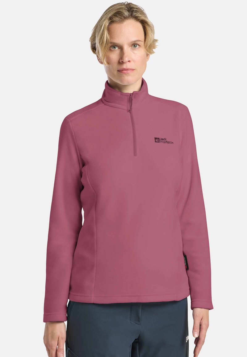 Donna con capelli biondi corti che indossa un maglione in pile a mezza zip color malva e pantaloni scuri, in piedi contro uno sfondo bianco semplice.