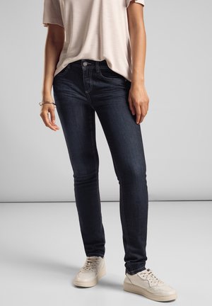 Jean skinny bleu foncé avec des coutures contrastées, taille moyenne et fermeture éclair. Porté avec une chemise claire et des baskets blanches.