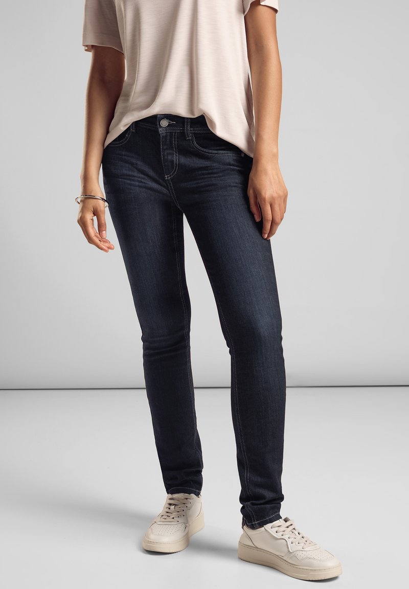 Tunna mörkblå jeans med kontrasterande sömmar, medellång midja och dragkedja. Bärs med en ljus skjorta och vita sneakers.