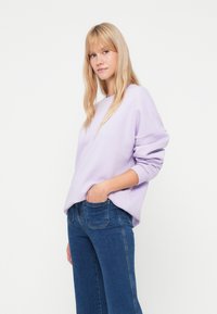 Ανοιχτό μωβ φούτερ με χαλαρή εφαρμογή, κατεβασμένους ώμους και ραμμένες μανσέτες, σε συνδυασμό με σκούρο μπλε παντελόνι με φαρδιά κουρτίνα. Λείο ύφασμα.