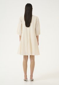 Cremefarbenes, kurzärmliges Kleid aus strukturiertem Stoff, mit ausgestelltem Rock und Ballonärmeln. Getragen mit hellen, mit Absatz versehenen Mules.
