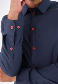 Camicia button-up blu navy con bottoni rossi, dotata di colletto classico e maniche lunghe. Il tessuto sembra liscio e strutturato per un aspetto aderente.