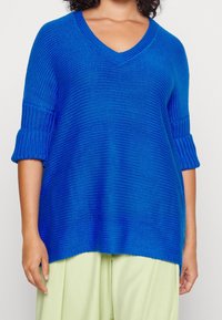 Blå stickad tröja med v-ringning, 3/4 ärmar och ribbade muddar. Mjuk textur och avslappnad passform, ihop med ljusgröna byxor.