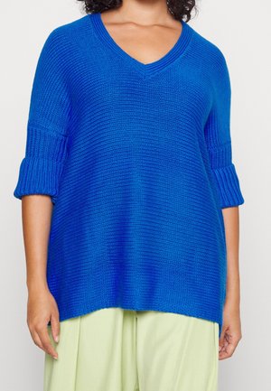 Blauer gestrickter Pullover mit V-Ausschnitt, 3/4-Ärmeln und gerippten Bündchen. Weicher Stoff und lockere Passform, kombiniert mit hellgrünen Hosen.