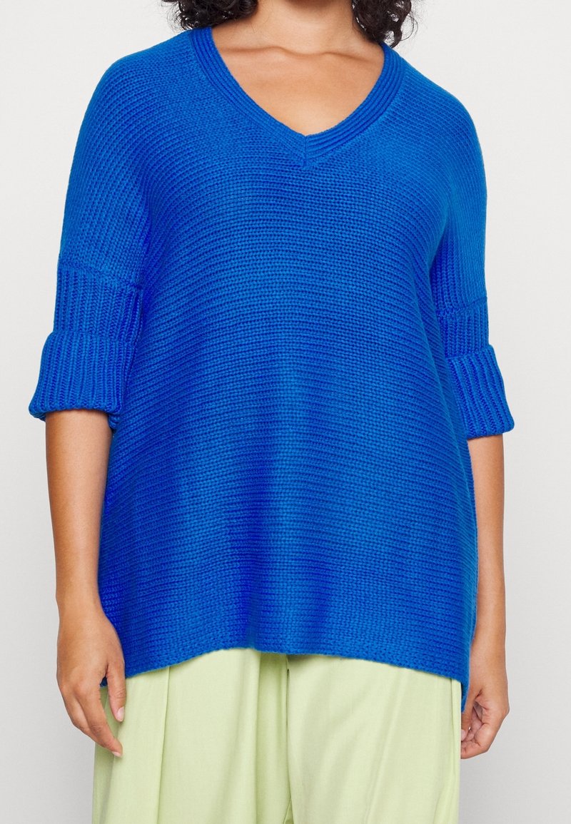 Blå stickad tröja med v-ringning, 3/4 ärmar och ribbade muddar. Mjuk textur och avslappnad passform, ihop med ljusgröna byxor.