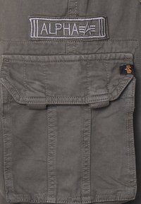 Olivenfargett stoffjakke med et brodert "ALPHA"-merke, en frontlomme og en kontrasterende oransje aksent på et merke.