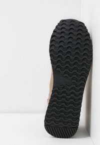 Suela de zapato con banda de goma negra que presenta patrones de olas y diagonales, parte superior beige parcialmente visible, sobre un fondo blanco en la esquina.