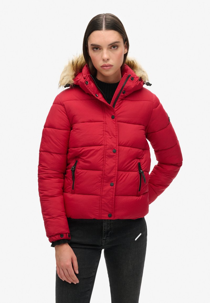 Giacca a vento rossa con cappuccio foderato in pelliccia sintetica, design a righe orizzontali, tasche con zip nere e chiusure a pressione. Indossata sopra un maglione nero.