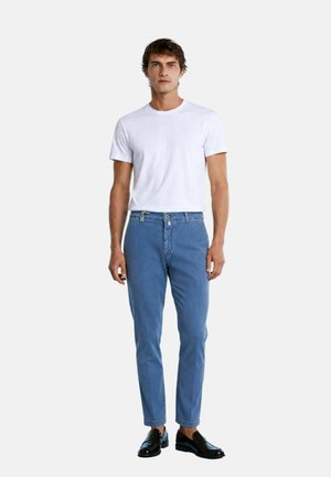 Uomo in piedi frontale che indossa una t-shirt bianca semplice, jeans blu slim fit e scarpe eleganti nere su uno sfondo chiaro uniforme.