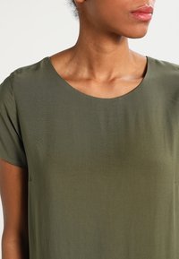 Haut vert olive à manches courtes avec une texture lisse et un col rond. Dispose de deux petites poches près de l'ourlet.