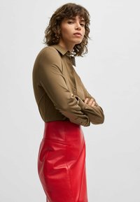 Camicia verde oliva a maniche lunghe con colletto e bottoni, abbinata a una gonna in pelle rossa a vita alta. La camicia ha una consistenza liscia.