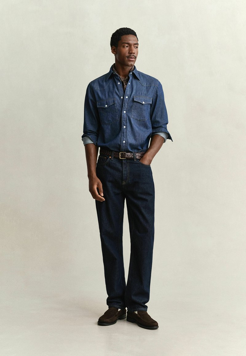 Man standing with one hand in pocket wearing a dark blue denim shirt, blue jeans, brown belt, and dark loafers against a plain background.